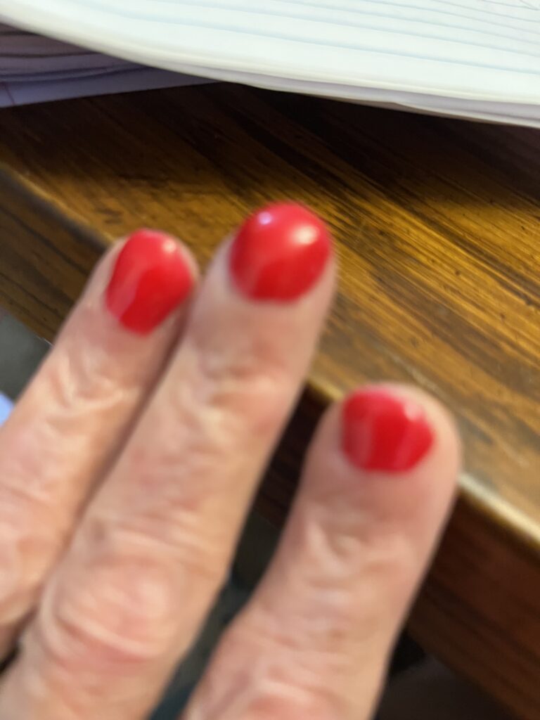 Close-up of two fingers with bright red nail polish resting on a wooden desk with papers in the background.